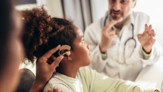 A young child watches her doctor as her mother tries to adjust her hearing aid while she is distracted. 